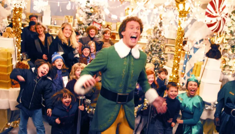 A foto mostra Will Ferrell em uma cena de 'Um Duende em Nova York'. Ele está cantando e várias crianças e adultos estão atrás dele, também cantando ou sorrindo.