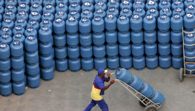 A foto mostra um homem empurrando quatro botijões azuis de gás. Diversos botijões parecidos estão empilhados ao fundo da foto.