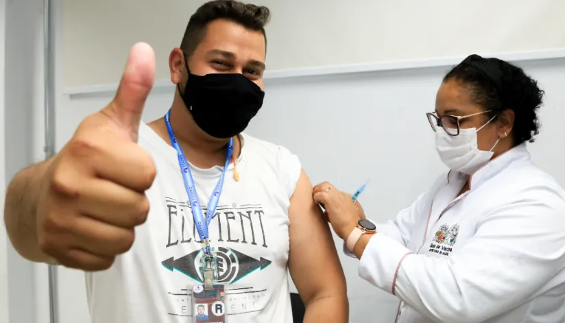 Homem vestindo uma camiseta branca e máscara preta faz sinal de joia enquanto enfermeira aplica vacina em seu braço esquerdo.