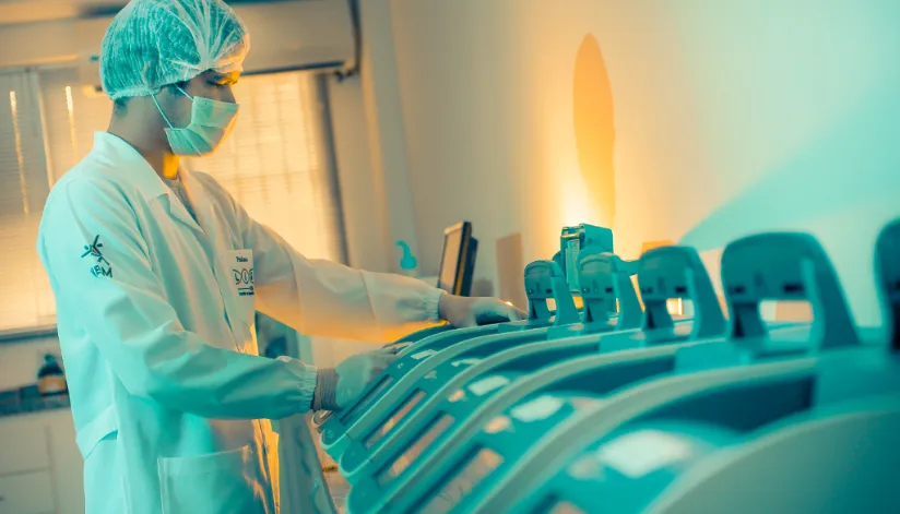 A foto mostra um homem vestido com um jaleco, máscara e touca em frente a aparelhos de laboratório.