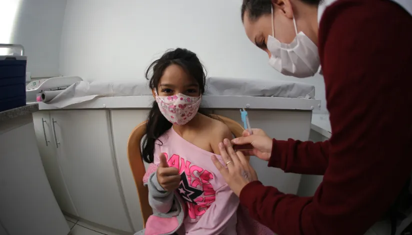 A foto mostra uma criança, que faz um sinal de positivo para a câmera, sendo vacinada por uma enfermeira.