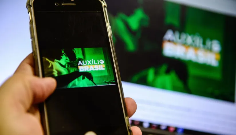 A foto mostra a tela de um celular com um banner do Auxílio Brasil. Ao fundo, é possível ver o mesmo banner na tela de um computador, desfocado.
