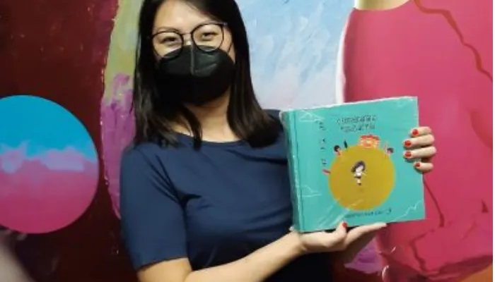 A autora Daniele Miki apresenta seu livro infantil para crianças surdas "O Extraordinário Mundo de Miki" em mãos, enquanto sorri por detrás da máscara de proteção. O livro possui capa azul clara com detalhes em amarelo.