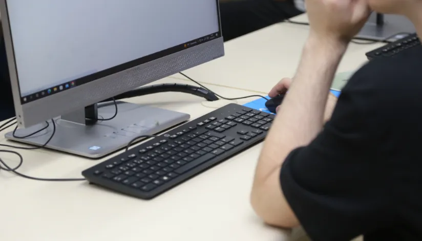 A foto mostra uma pessoa em frente a um computador. Ele está vestido de preto, com o cotovelo esquerdo em cima da mesa.