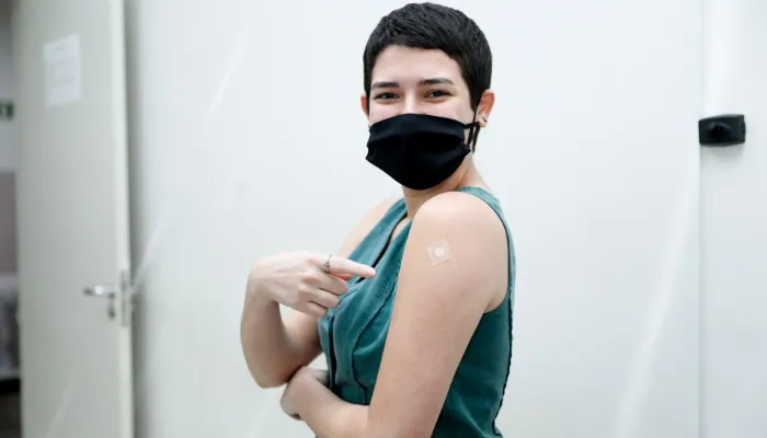 A imagem mostra uma mulher apontando para seu braço esquerdo, onde está um curativo da vacina.