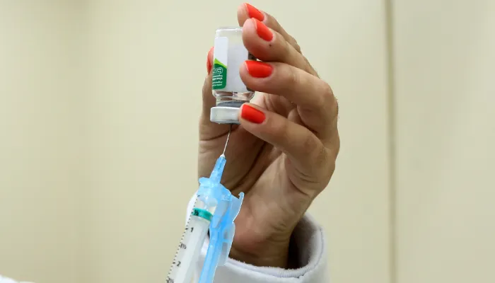 Na imagem, a fotografia apresenta uma mão feminina com esmalte laranja enchendo uma seringa com um dose de vacina.