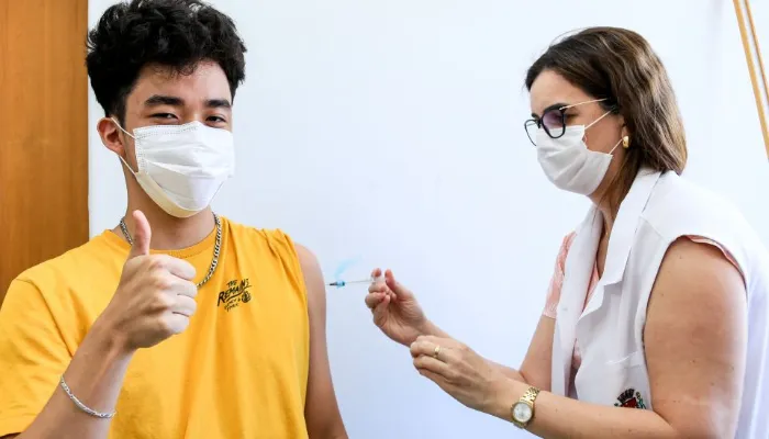 Jovem de camiseta amarela recebendo vacina no braço enquanto faz o sinal positivo para a câmera.