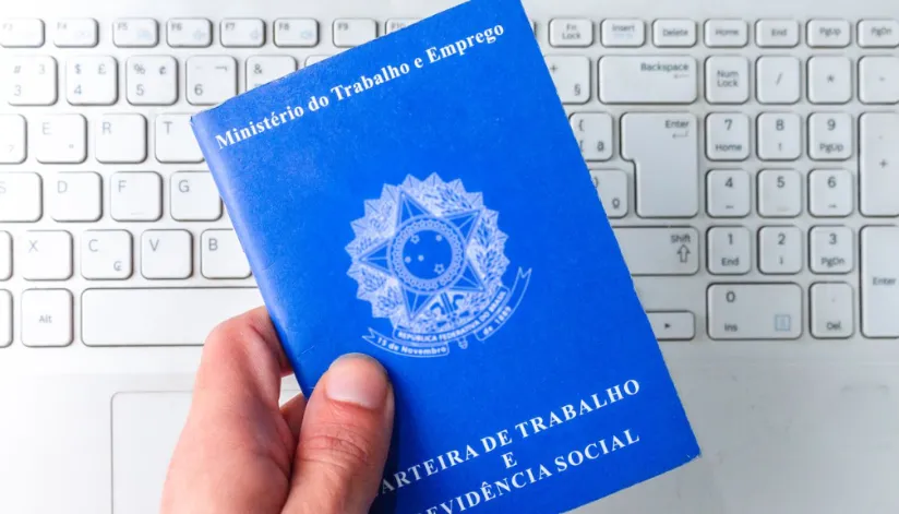 Na imagem, uma mão segura a carteira de trabalho acima de um teclado de computador branco. A carteira azul cobalto apresenta o brasão do Ministério do Trabalho.
