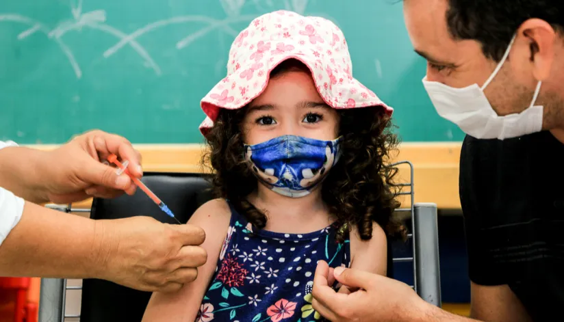 Criança de cabelos cacheados recebe vacina no braço. Mesmo de máscara, é possível notar que a crinça sorri, enquanto segura a mão de seu pai.
