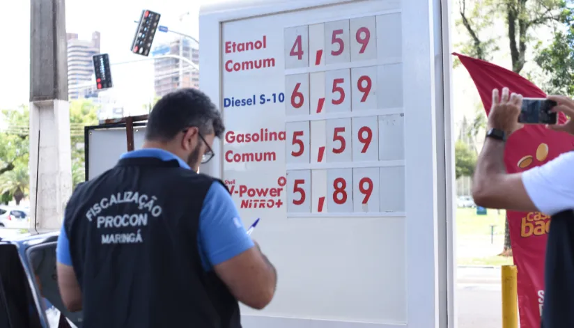 A foto mostra um agente de Procon em frente a uma placa com o preço dos combustíveis.