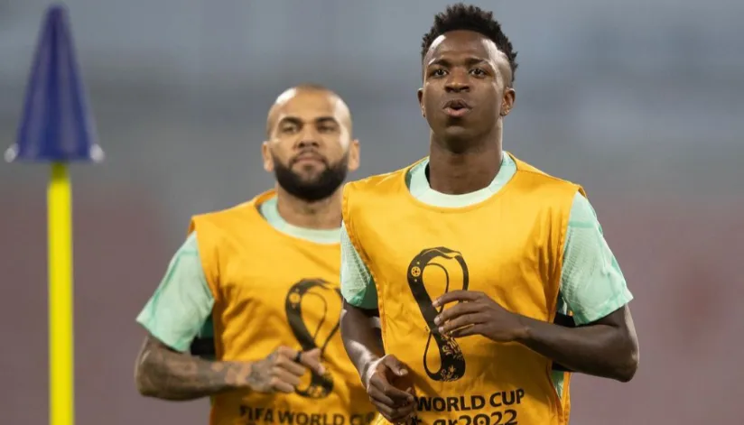 A foto mostra o jogados Vinicius Jr. em primeiro plano e Daniel Alves logo atrás. Ambos estão vestidos com camisetas verde claro e com um colete amarelo, com o símbolo da copa do mundo, por cima.