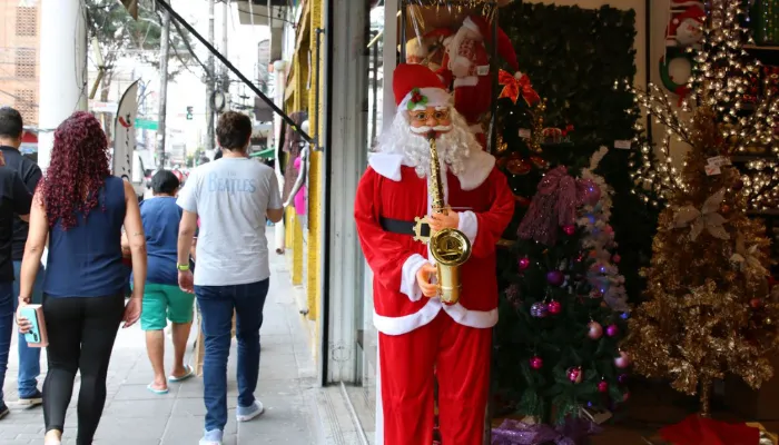 A foto mostra uma rua de comércio. Na porta de uma loja, do lado direito da imagem, há um papai noel e, do lado esquerdo, pessoas andam na calçada.