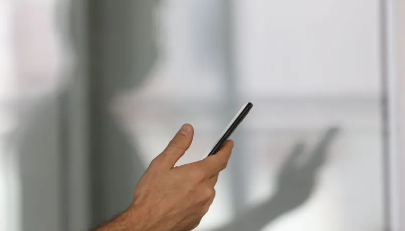 A foto mostra uma mão segurando um celular. Atrás, é possível ver o reflexo da pessoa em um vidro.