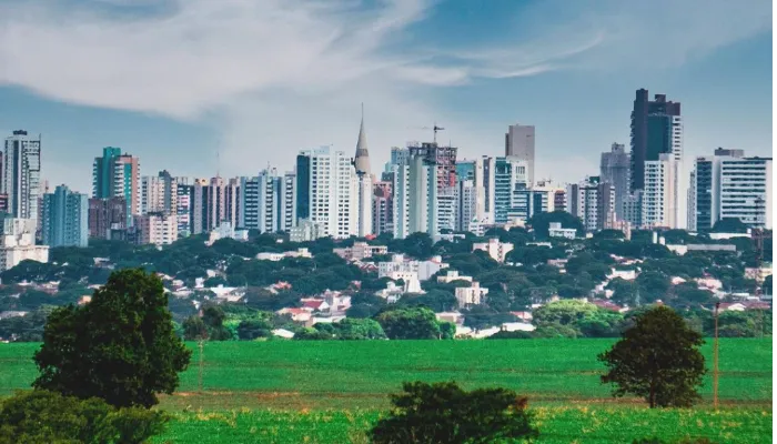 A foto mostra o panorama de Maringá, visto de longe. No primeiro plano da foto, há árvores e um grande gramado verde enquanto, ao longe, estão as casas e prédios da cidade.