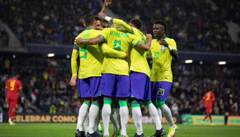 A imagem mostra os jogadores da seleção brasileira se abraçando em círculo. Todos estão de costas para a câmera, com exceção do camisa 20, Vinícius Junior.