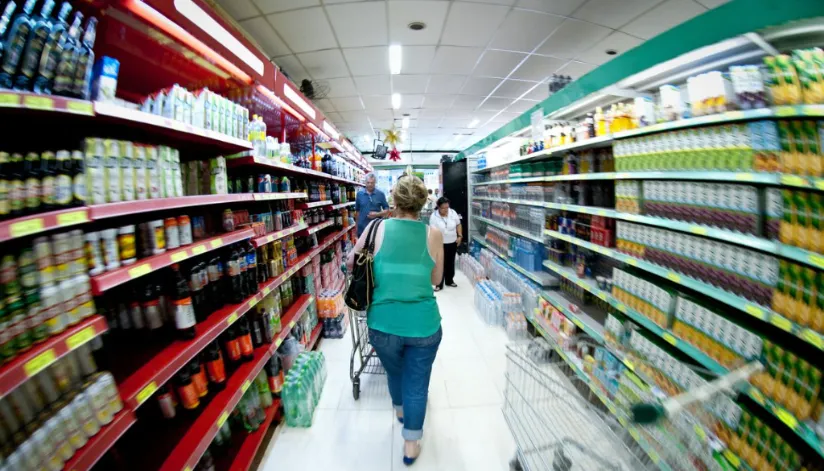 A foto mostra uma mulher de costas, empurrando um carrinho em um corredor de supermercado.