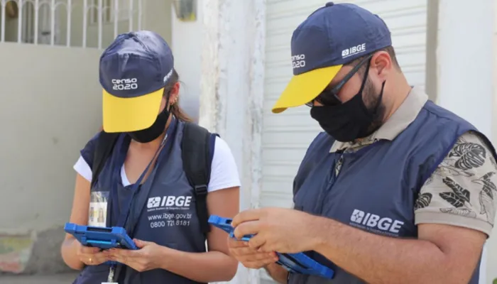 A foto mostra dois agentes do IBGE olhando para baixo enquanto seguram tablets em suas mãos. Ambos usam um colete com o logo e informações do IBGE e bonés escrito "Censo 2020"