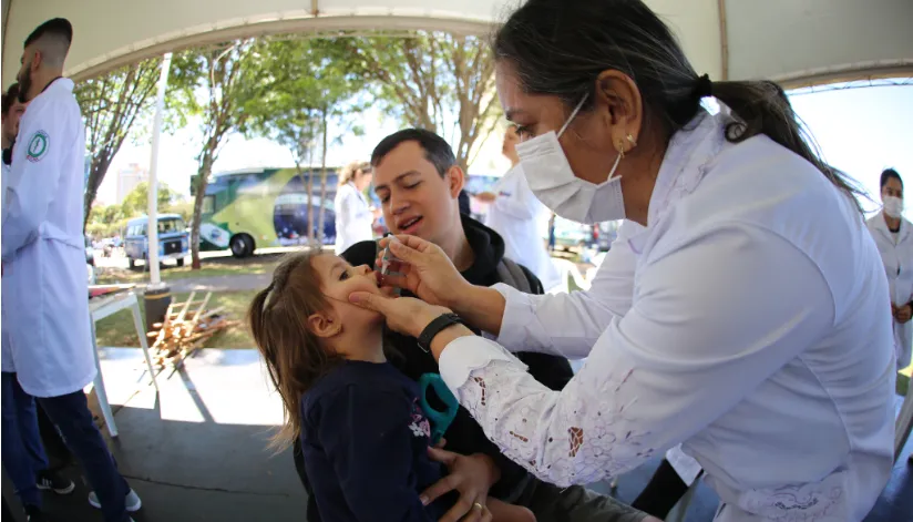 A foto mostra uma agente de saúde, embaixo de uma tenda, dando uma gotinha de vacina para uma menina, que está sentada no colo de um homem. Ao fundo, é possível ver outros agentes de saúde