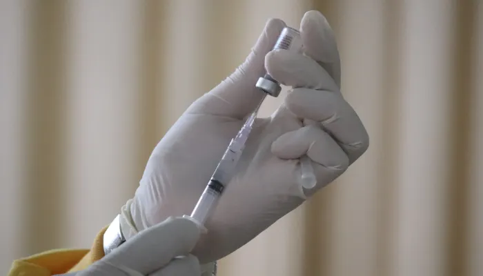 A foto mostra uma mão segurando um frasco de vacina de cabeça para baixo, enquanto a outra mão segura uma seringa, aspirando o líquido