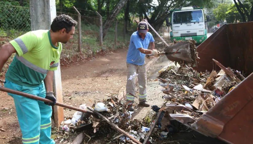 A imagem mostra dois servidores da prefeitura de Maringá recolhendo o lixo e o colocando em uma caçamba.