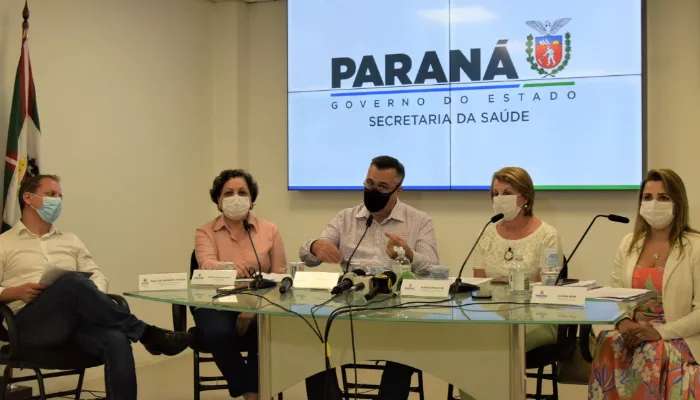 A foto mostra cinco pessoas sentadas em volta de uma mesa, em uma coletiva de imprensa. Microfones estão postados em frente de quatro delas e o secretário de saúde Beto Preto está sentado no meio delas.
