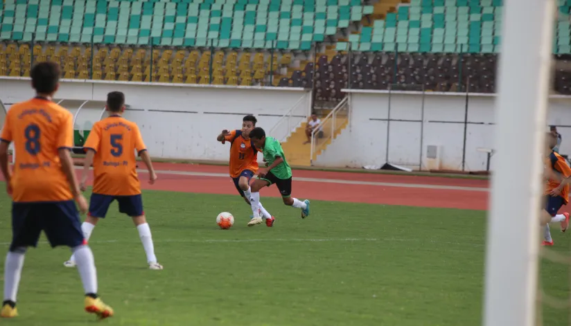 A imagem mostra quatro meninos em um campo de futebol. Um deles, vestido de laranja, corre atrás da bola enquanto um menino vestido de verde também a disputa.