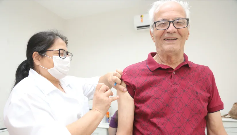A foto mostra uma enfermeira aplicando a vacina no braço de um senhor, que está sorrindo para a câmera.