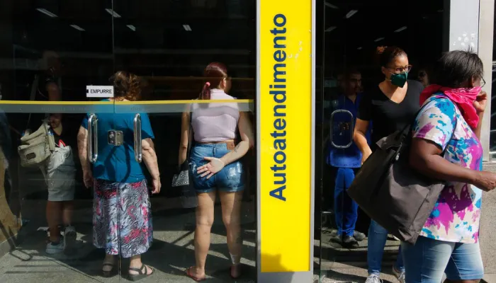 A foto mostra pessoas em uma agência do Banco do Brasil. Do lado esquerdo da imagem, é possível ver pessoas em pé, dentro da agência e, do lado direito, algumas pessoas se dirigem para fora.