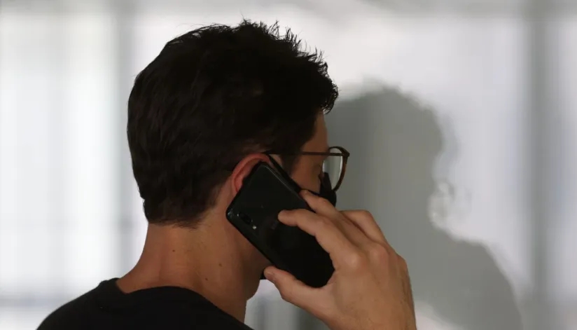 Homem atende o celular de costas para a câmera. Ele usa um óculos de grau e sua sombra é projetada na parede em sua frente.