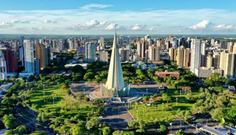 Foto aérea de Maringá na região da Praça da Catedral em um dia ensolarado na cidade. O verde das árvores e do gramado da praça se destacam em meio à arquitetura acinzentada e o céu azul claro intenso.