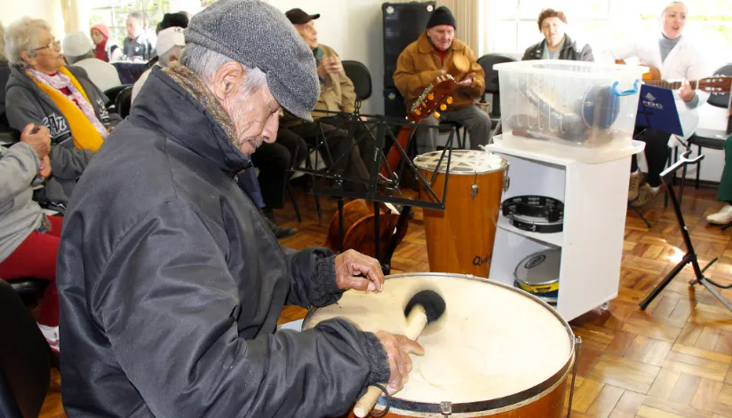A foto mostra um idoso tocando um tambor. Ao fundo é possível outros idosos tocando instrumentos.