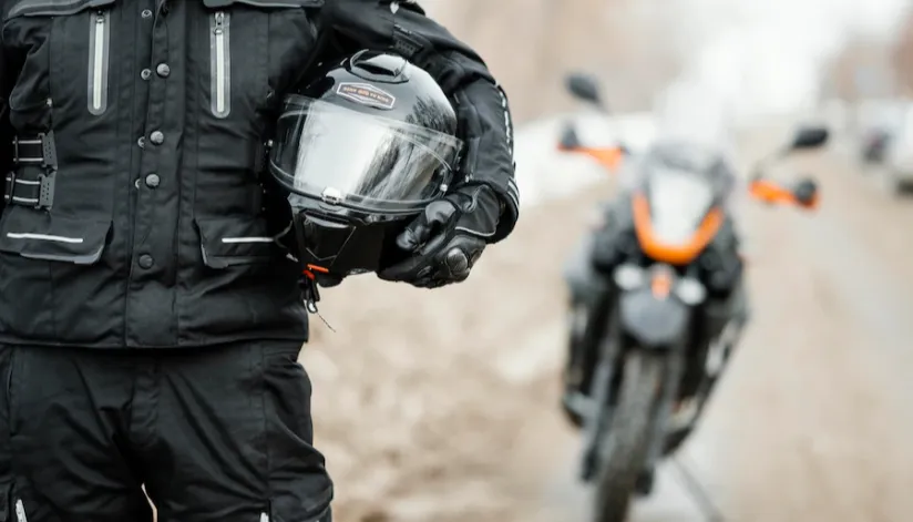 Motociclista segura o capacete abaixo de seu braço em primeiro plano. Desfocada, ao fundo está a moto em uma estrada.