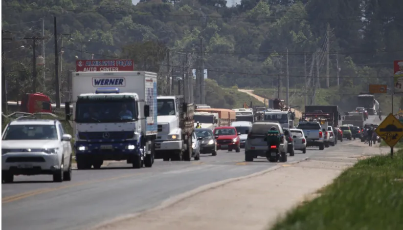 A foto mostra uma estrada com um congestionamento de carros e caminhões do lado esquerdo da imagem e veículos andando na direção contrária do lado direito.