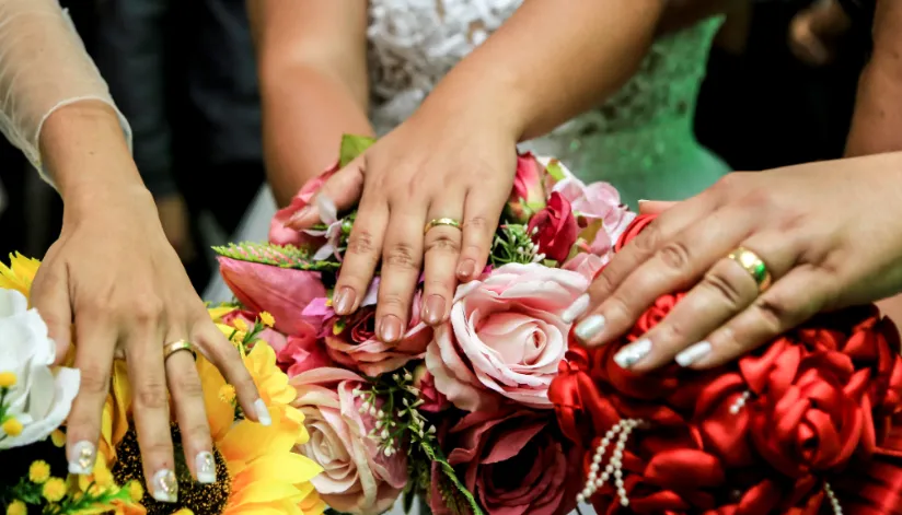 Três noivas mostram a mão com anel de ouro sob o buquê de cada uma. São buquês amarelo, rosa e vermelho.