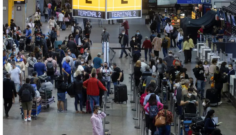 A foto mostra um terminal de aeroporto com diversos passageiros enfileirados e andando pelo terminal.