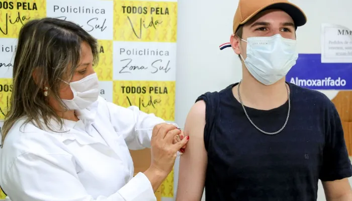 Jovem de boné marrom, camiseta preta e máscara de proteção branca recebendo a vacina no braço.