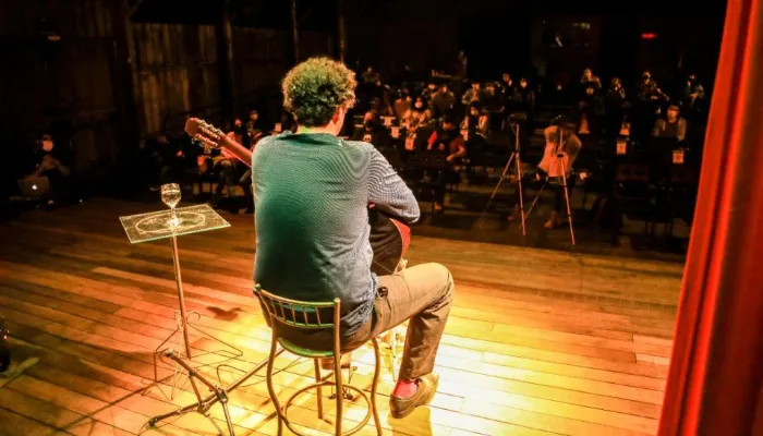 Fotografia tirada de um teatro cheio de pessoas assistindo à apresentação de um músico sentado com o violão em mãos.