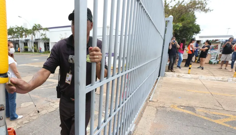 A foto mostra um segurança fechando o portão de uma escola.