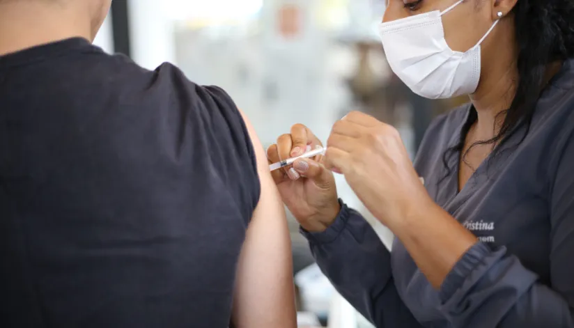 A foto mostra uma enfermeira vacinando uma pessoa. A pessoa vacinada está de costas para a câmera e a enfermeira usa uma máscara.