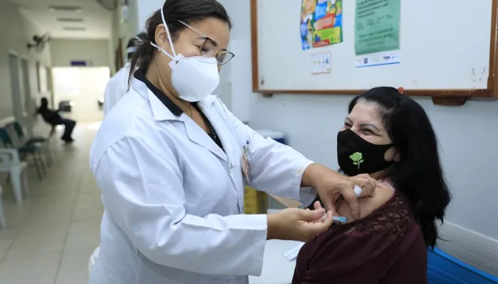 Na foto, uma enfermeira aplica a vacina no braço de uma mulher sentada, as duas estão de máscaras mas aparentam estar sorrindo.