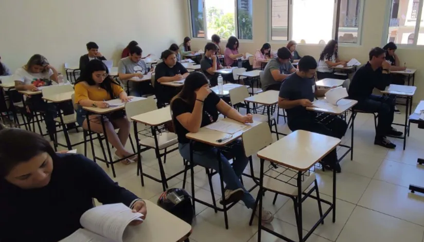 A foto mostra uma sala de aula com vestibulandos sentados, fazendo a prova.