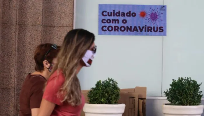 Imgem de dois pedestres mascarados passando em frente a um cartaz com o aviso "cuidado com o coronavírus".