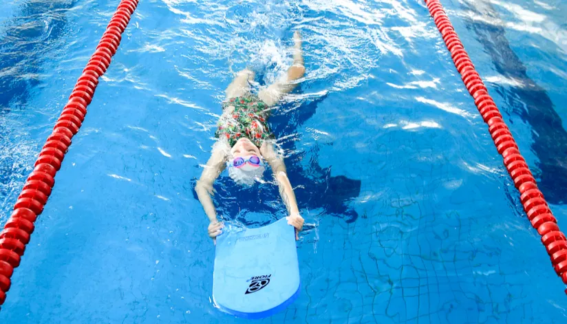 A foto mostra uma menina boiando em uma piscina olímpica, segurando uma pequena prancha